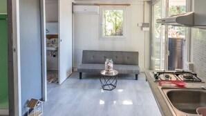 A compact kitchen with a grey sofa, a small table with a glass jar, and a window with a view of greenery. - Agricampeggio Princess - (3) (Fiorentina)