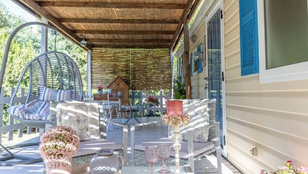 Een overdekt terras met rieten meubilair, een glazen tafel met roze bloemen, en een huis met blauwe luiken.
