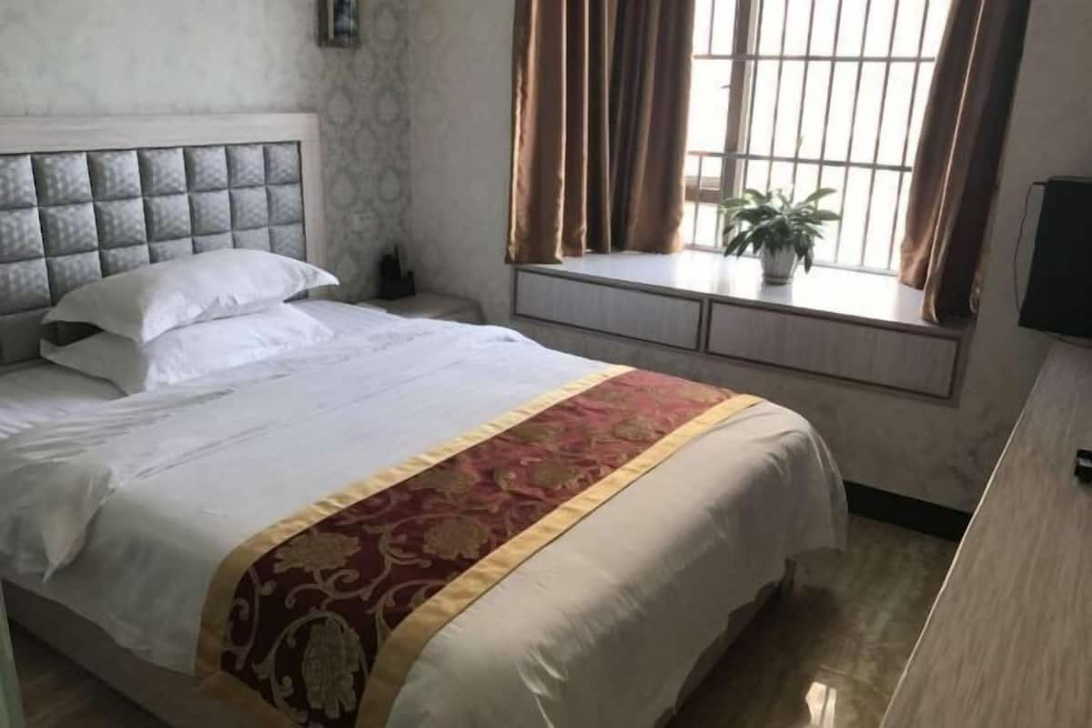 Een netjes opgemaakt bed met hoofdbord, een nachtkastje met een plant en een raam met gordijnen.