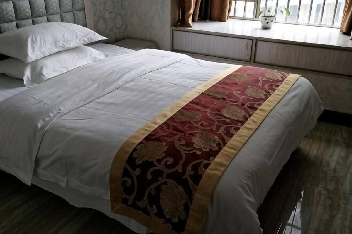 Een net opgemaakt bed met een wit-rode spreideken, een raam met gordijnen en een nachtkastje met een lamp.