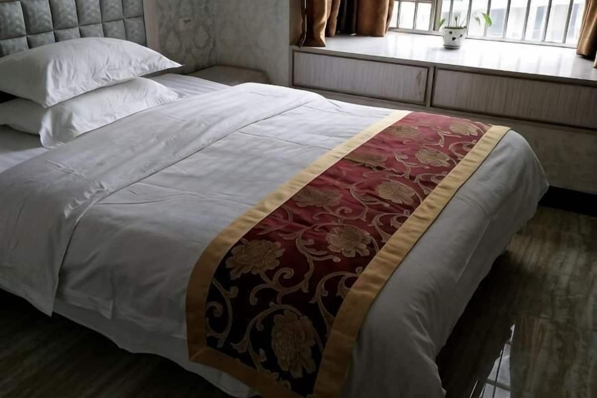 Una cama bien hecha con una colcha blanca y roja, una ventana con cortinas y una mesita de noche con una lámpara.