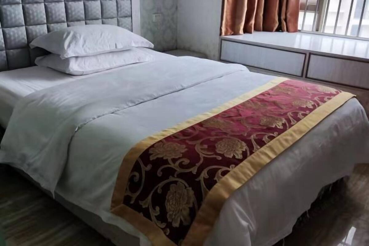 Een net opgemaakt bed met een bedsprei met patroon, een gestoffeerd hoofdbord en een raam met gordijnen.