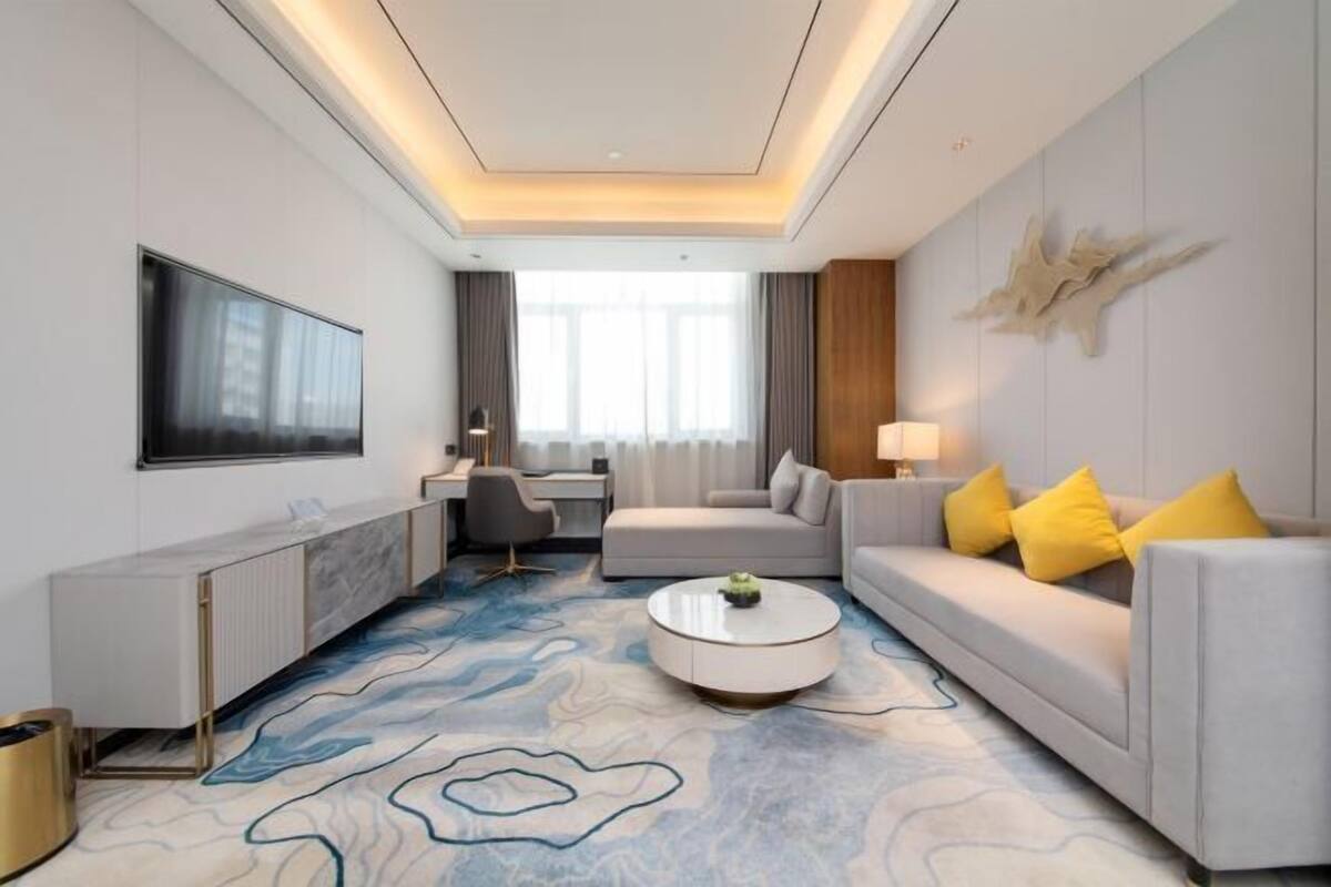 Une chambre d’hôtel moderne dotée d’un grand téléviseur à écran plat, d’un canapé avec des coussins jaunes, d’une table basse ronde et d’un tapis à motifs.