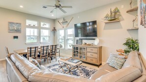 A living room with a beige sofa, a coffee table, a TV, a dining area with chairs and a table, and a ceiling fan. - Walk to Sea Isle Beach! Canal-front Gem w/ Pool (Galveston)