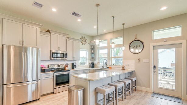 A modern kitchen with stainless steel appliances, a large island with bar stools, and a view to the outside. - Walk to Sea Isle Beach! Canal-front Gem w/ Pool (Galveston)
