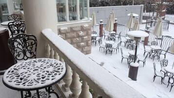 En snötäckt uteplats med runda bord och stolar, en stenmur och utsikt över en snöig gata.