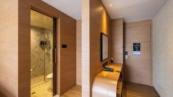 A modern hotel room with a wooden floor, a glass shower enclosure, a wooden bench, and a wall-mounted TV.