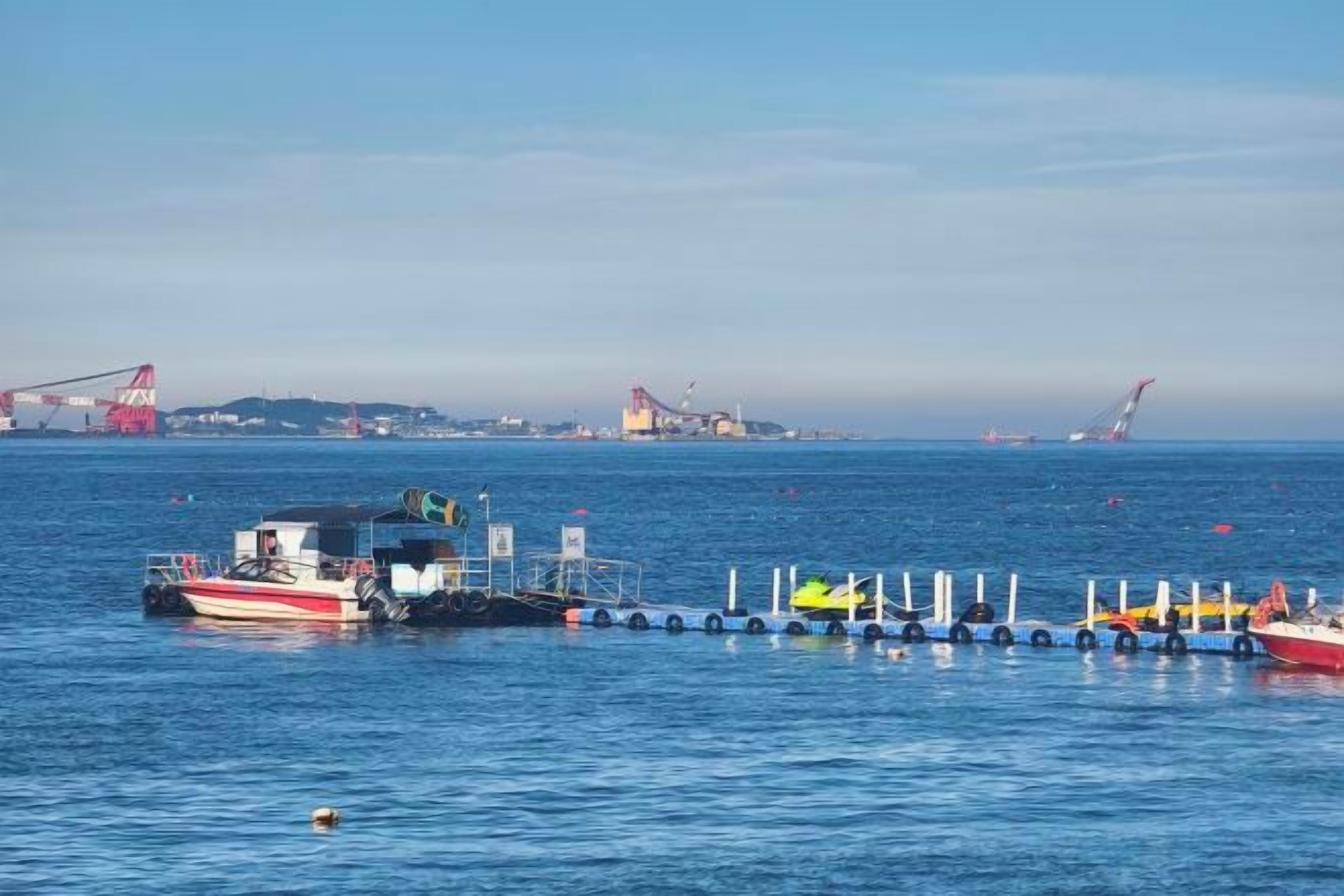 A calm body of water with a dock and boats, a clear sky, and distant industrial structures.