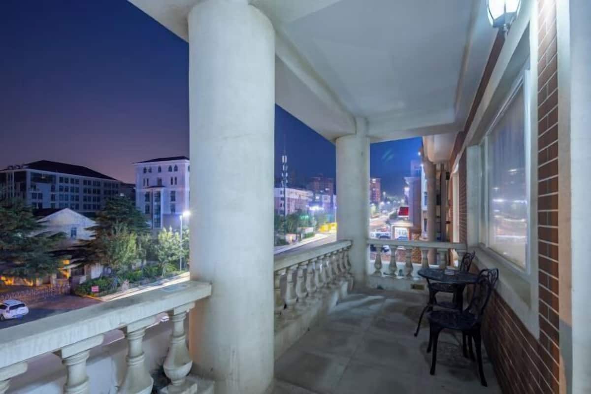 A balcony with a view of a cityscape at dusk, featuring a railing, a table with chairs, and a building with large windows.