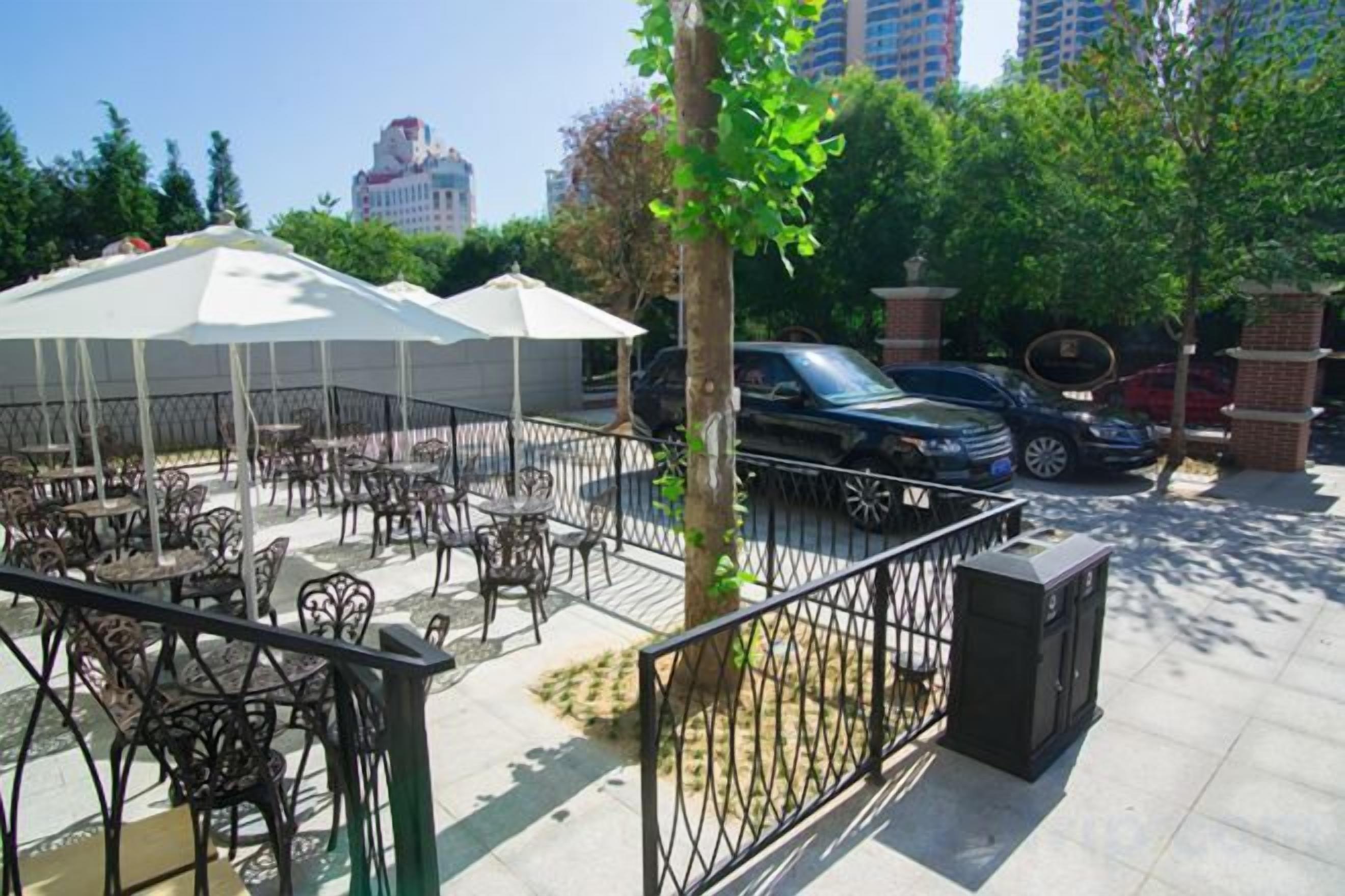 An outdoor cafe with tables and umbrellas, a tree, and parked cars.