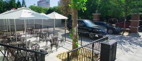 An outdoor cafe with tables and umbrellas, a tree, and parked cars.