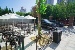 An outdoor cafe with tables and umbrellas, a tree, and parked cars.