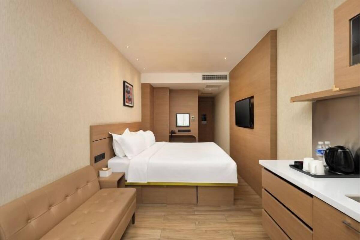 A hotel room with a bed, bedside table, a bench, a TV, and a kitchenette area.