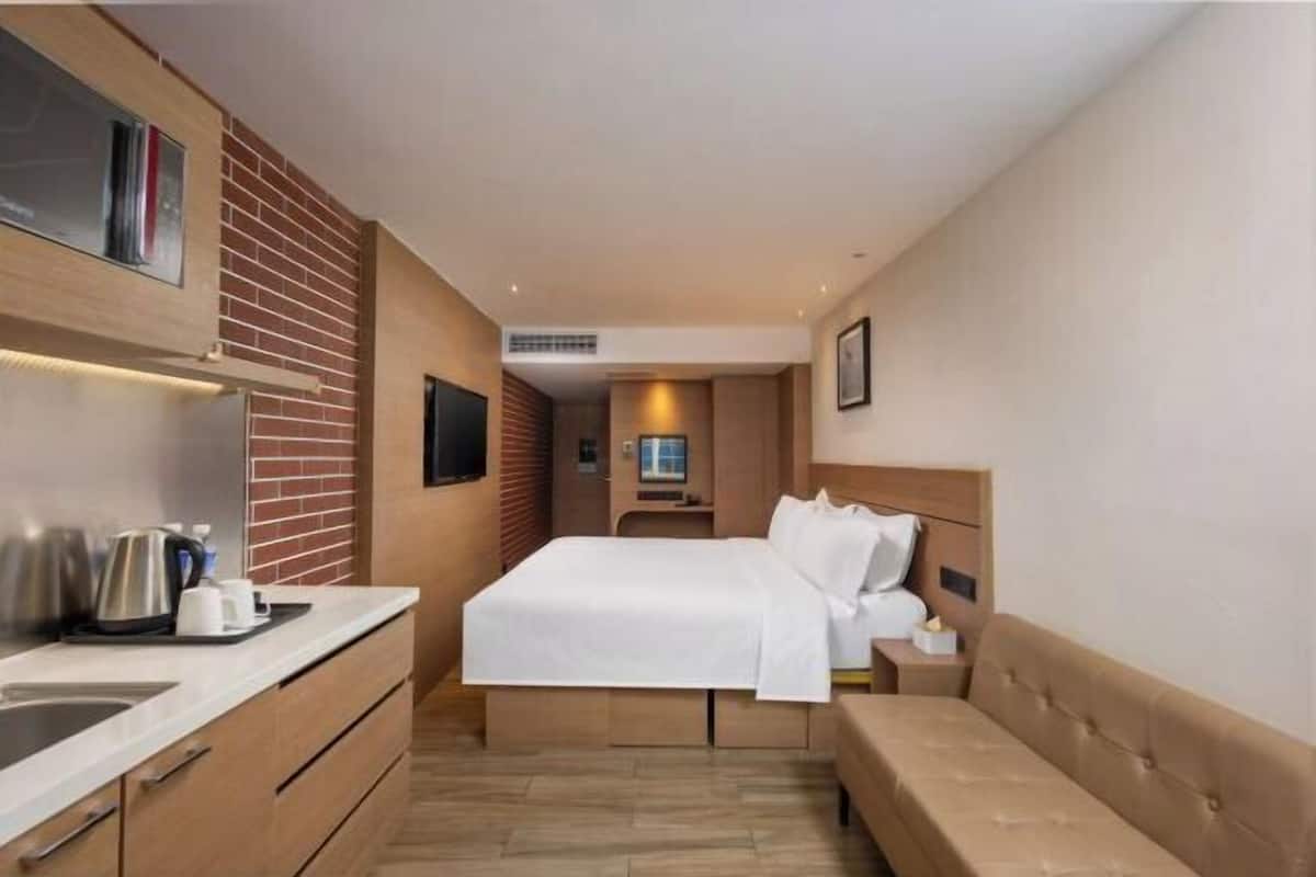 A modern hotel room with a bed, bedside table, and a kitchenette area.