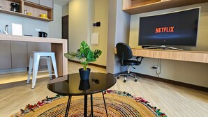 A modern living room with a flat-screen TV displaying 'NETFLIX', a small white stool, a black office chair, a glass coffee table with a plant, and a colorful rug. - Exclusive modern apartment with unbeatable location (Cochabamba)