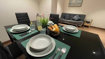 Uma área de refeições com mesa preta, jogos americanos verdes, pratos brancos e talheres. Há uma cesta de pão e um vaso com flores sobre a mesa.