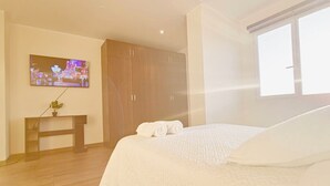 A bedroom with a bed, a TV mounted on the wall, a wooden wardrobe, and a small table with a plant. - Large and spacious apartment, well equipped and located (Cochabamba)