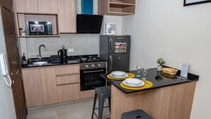 Cozinha compacta com fogão a gás, forno, micro-ondas e frigorífico. Possui dois taburetes e um pequeno espaço de refeições com pratos e copos.