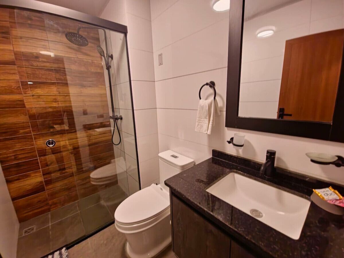 Un baño moderno con ducha de vidrio, inodoro blanco y lavamanos con encimera oscura.