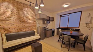 Ein modernes Wohnzimmer mit Backsteinwand, einer Couch, einem Esstisch mit Stühlen und einer Küchenzeile inklusive Mikrowelle und Kühlschrank.