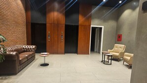 A lobby with a brown leather sofa, a beige armchair, and a small round table. There are two elevators with digital displays.