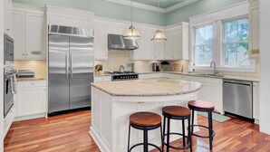A kitchen with white cabinets, a stainless steel refrigerator, a gas stove, and a large island with stools. - POOL, ROOF DECK, ELEVATOR, 5 MIN TO THE SURF! (Isle of Palms)
