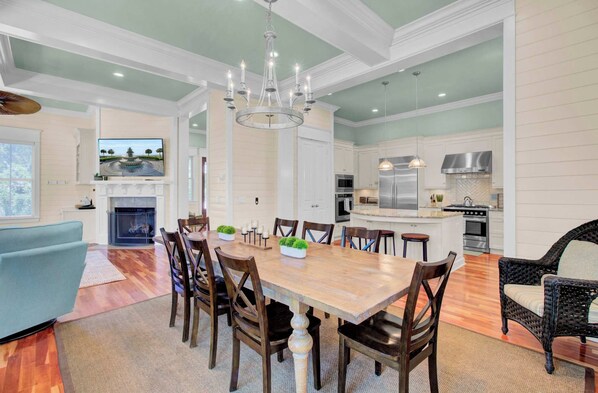 A spacious dining area with a wooden table and chairs, a chandelier, and a television mounted on the wall.