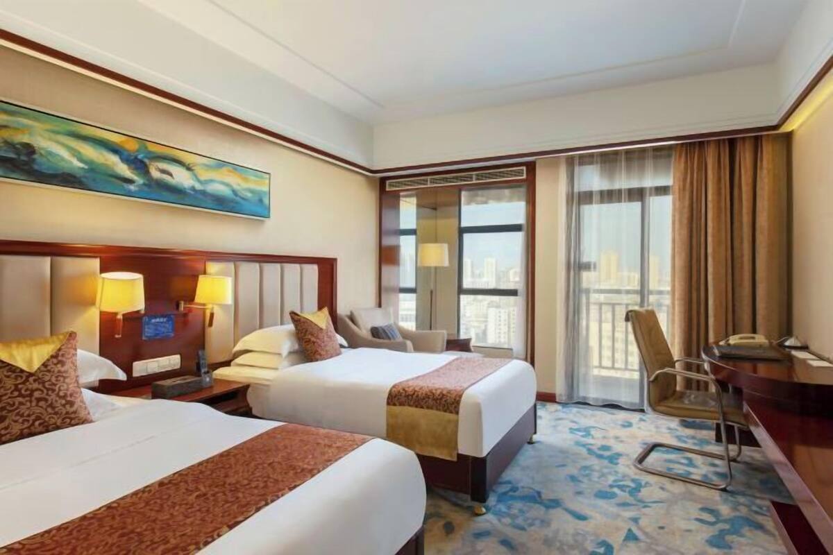 Ein Hotelzimmer mit zwei Betten, einem Schreibtisch, einem Stuhl und einem großen Fenster mit Blick auf die Stadt.