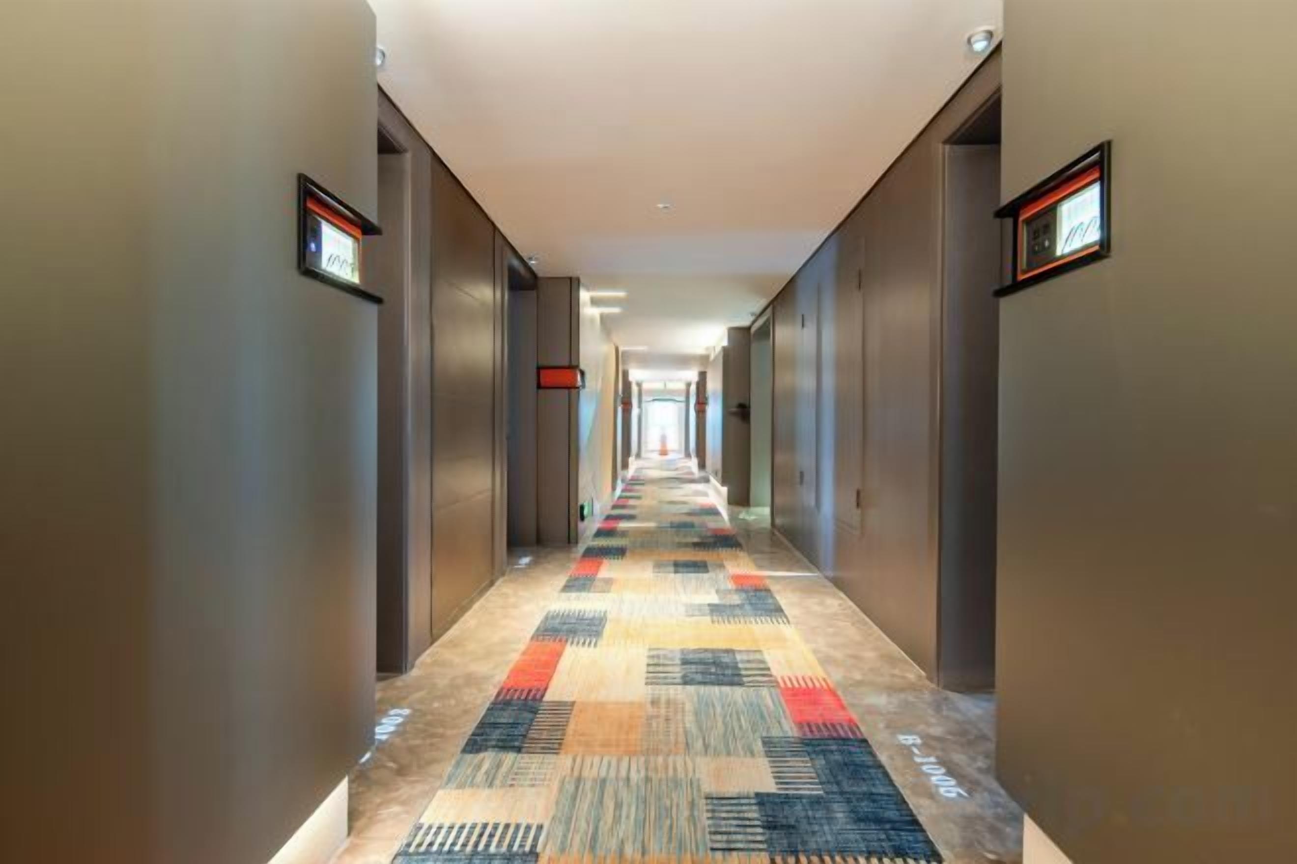 A hotel corridor with carpeted flooring, doors on both sides, and a long hallway leading to an elevator.