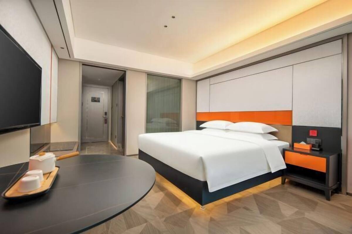 Ein Hotelzimmer mit Bett, Fernseher, Schreibtisch und einer Ablage mit Gegenständen, sowie einer Glastür zum Badezimmer.