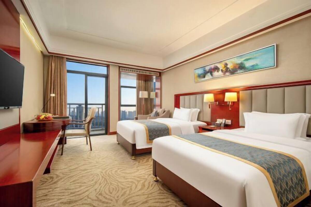 Ein Hotelzimmer mit zwei Betten, einem Schreibtisch, einem Stuhl, einem Fernseher und einem großen Fenster mit Blick auf die Stadt.