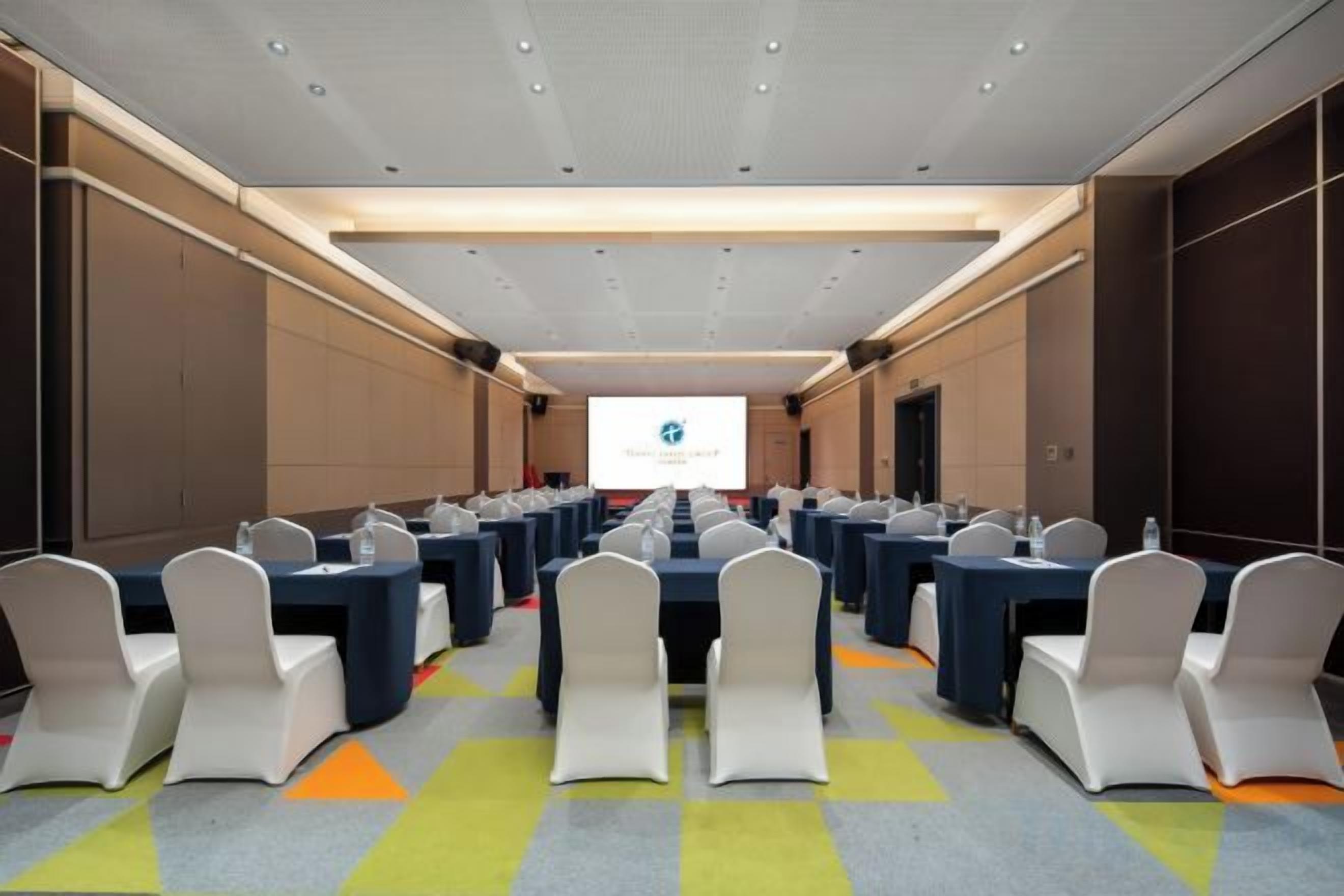 A conference room with rows of tables and chairs, a presentation screen, and a carpeted floor.