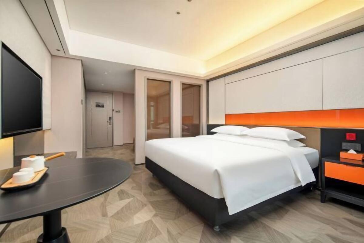 Ein Hotelzimmer mit einem Bett, einem Fernseher, einem Schreibtisch mit einer Tablett voller Tassen und einer Tür zu einem anderen Raum.