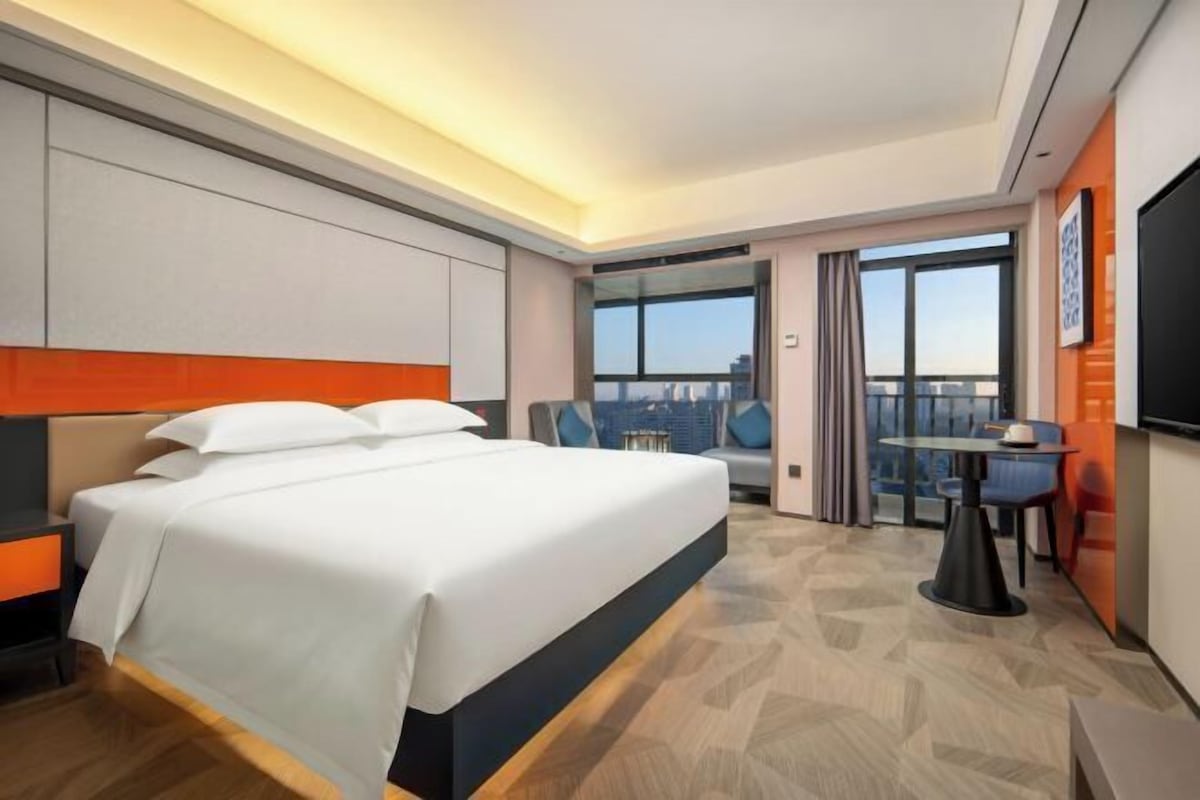 Habitación de hotel con una cama grande, un escritorio, una silla, un televisor y un balcón con vistas a la ciudad.