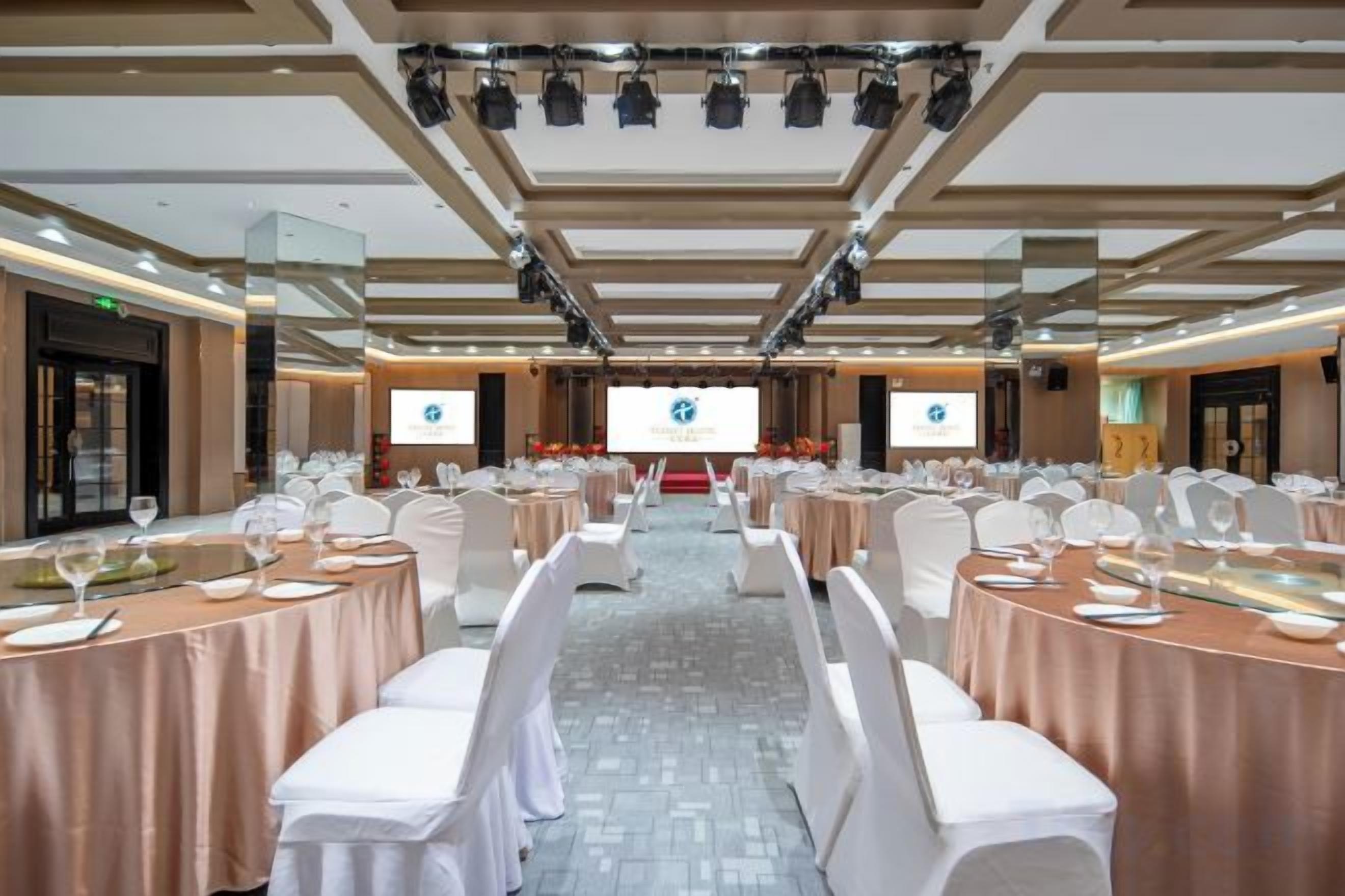 Un salón de conferencias con mesas redondas, sillas blancas y un escenario con una pantalla de presentación.