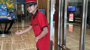 A person in a red uniform standing in a lobby with a glass partition, a digital screen, and a table with a plant.