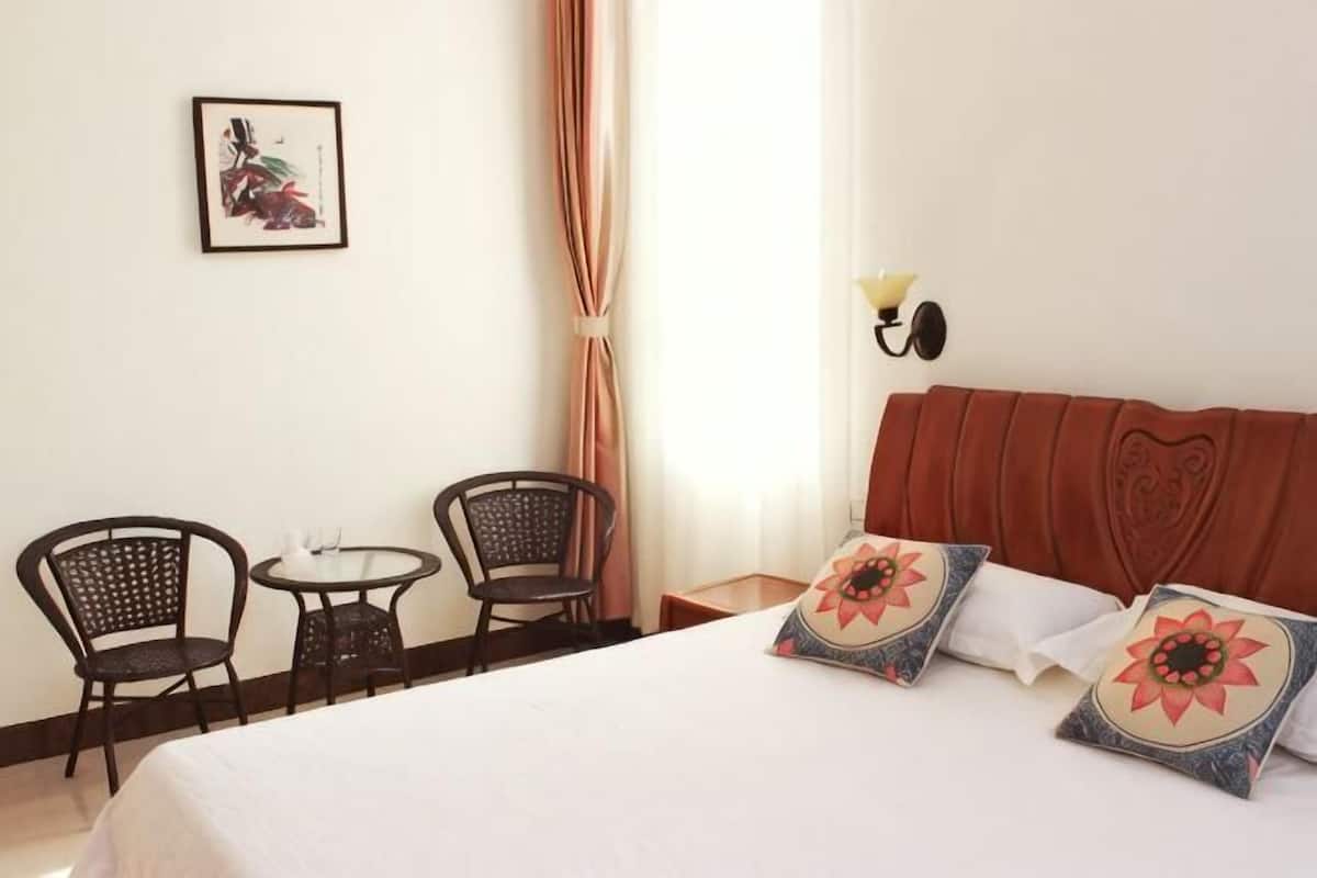 A hotel room with a bed, two chairs, a small table, a wall-mounted lamp, and a framed picture.