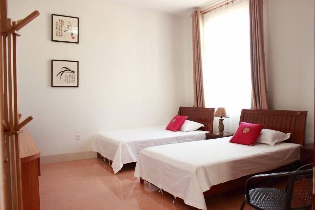 A room with two single beds, a wooden chair, a nightstand with a lamp, and two framed artworks on the wall.