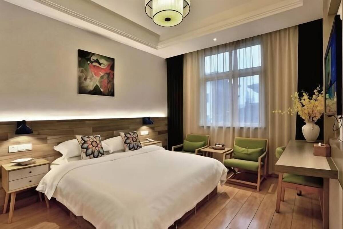 A hotel room with a large bed, bedside tables, a green armchair, a small table, and a window with curtains.