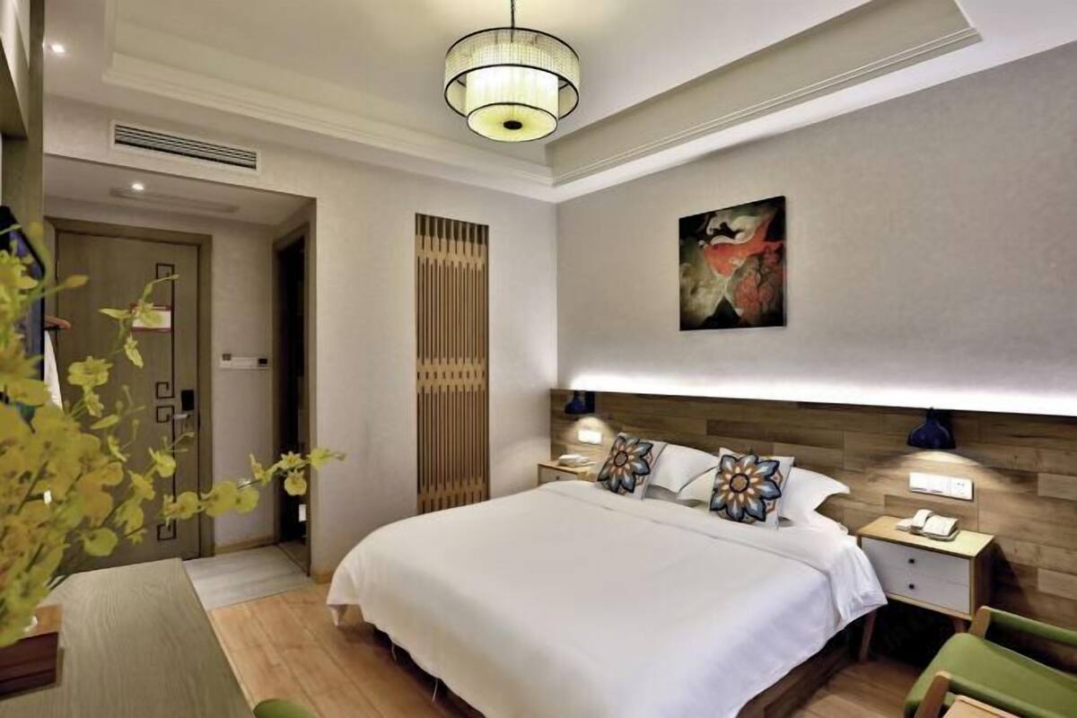 A modern hotel room with a large bed, bedside lamps, a wooden headboard, a painting on the wall, and a potted plant.