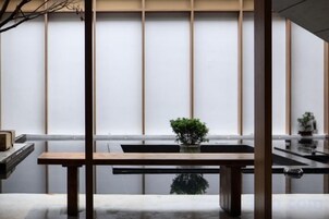 Una habitación con un banco de madera, una planta pequeña en maceta y vistas a través de una ventana con persianas verticales.