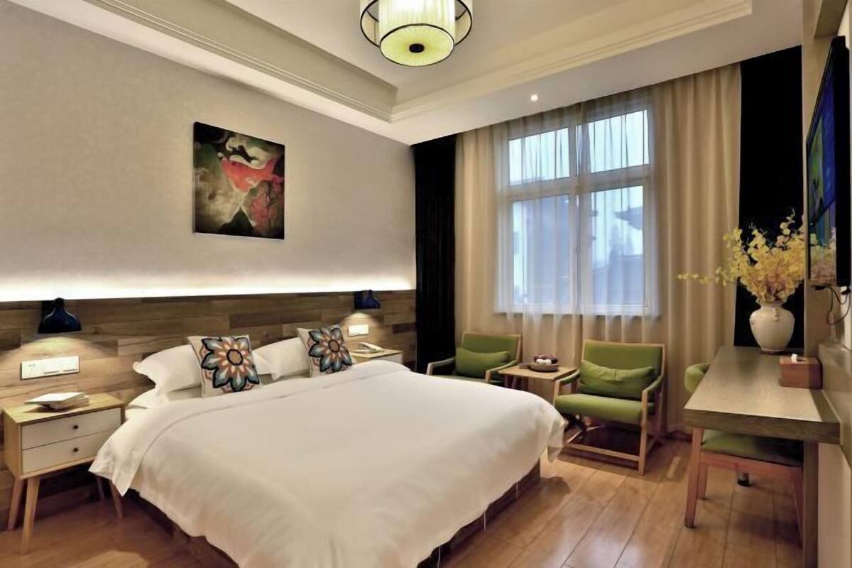 A hotel room with a large bed, a desk, a chair, a nightstand, a lamp, a painting on the wall, and a window with curtains.