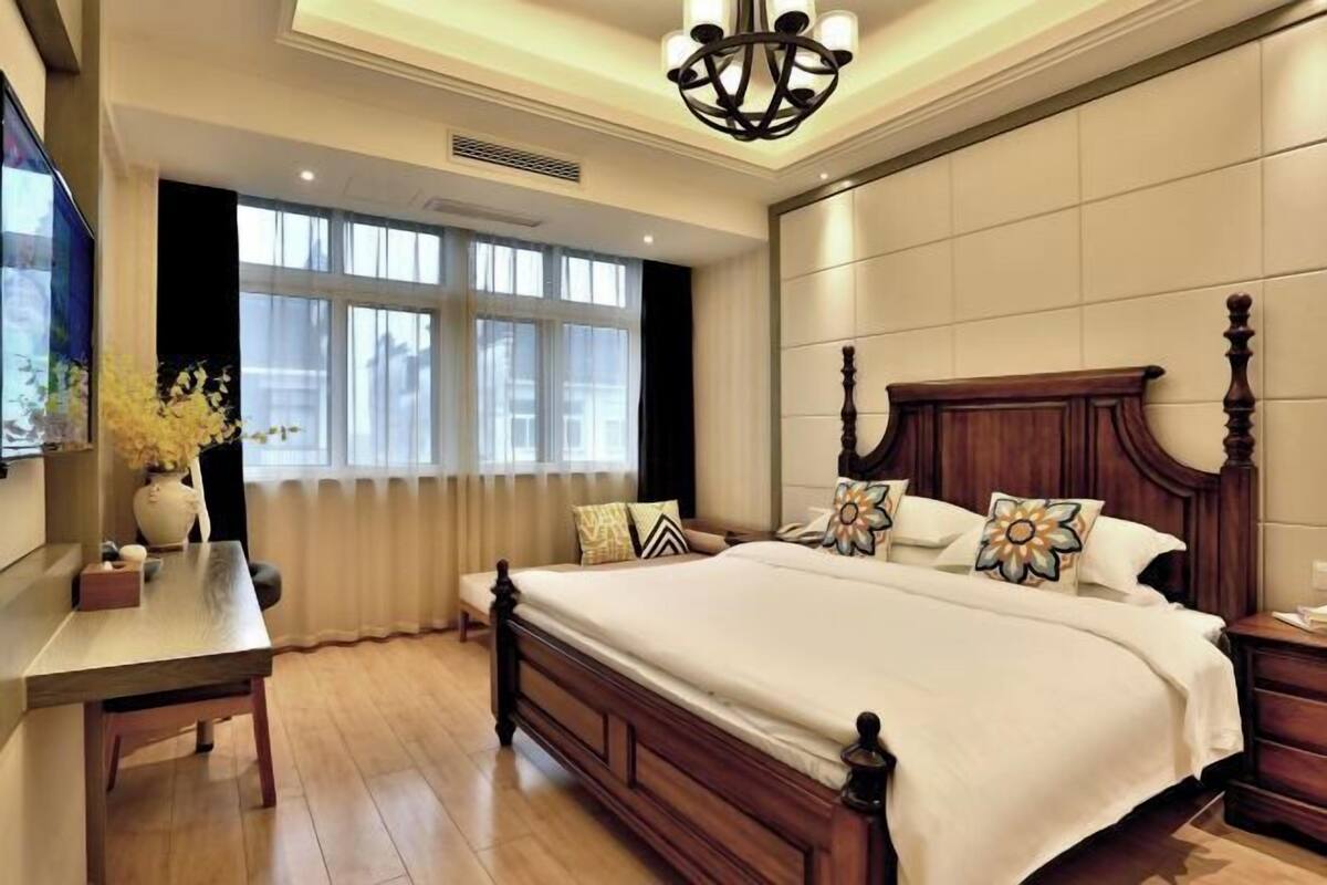 A bedroom with a large bed, a bedside table, a chair, a desk, and a window with curtains.