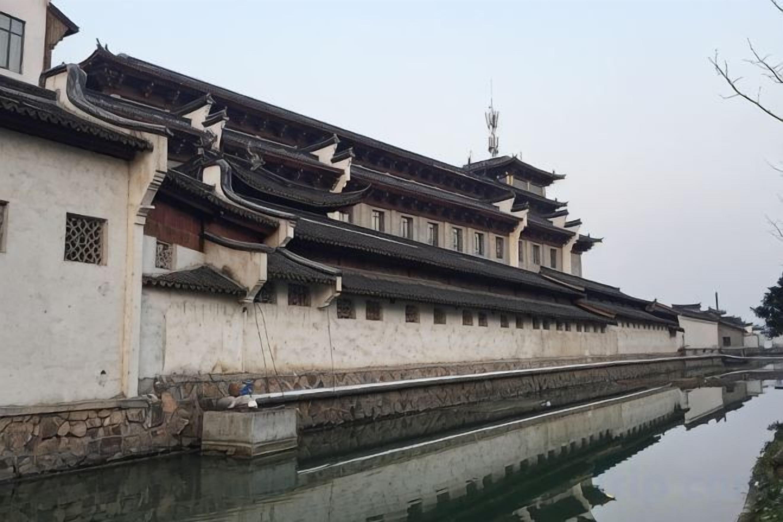 A traditional building with a long, narrow structure, a stone wall, and a waterway in front.