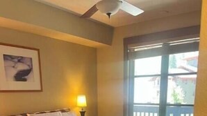 A hotel room with a bed, a ceiling fan, a window with a view of buildings, and a framed picture on the wall.