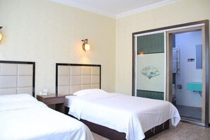 Ein Hotelzimmer mit zwei Betten, einem Nachttisch, einer Wandleuchte und einem durch eine Glastür einsehbaren Badezimmer.