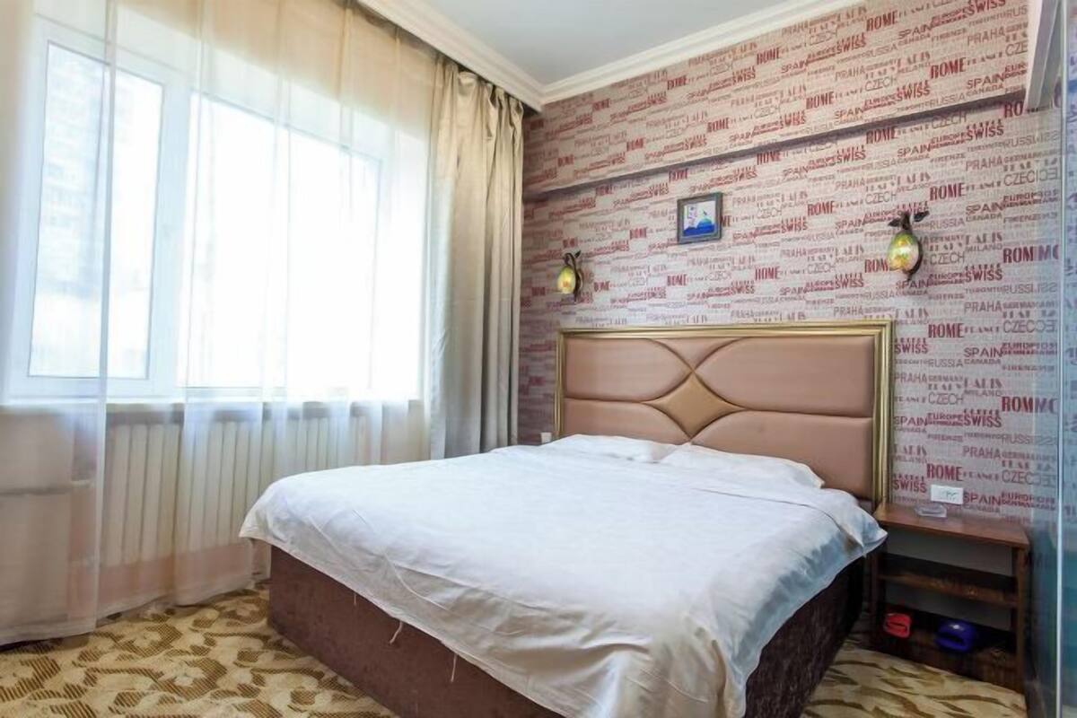 Een hotelkamer met een bed, een raam met gordijnen, een radiator, een muur met tekst erop, en twee wandlampen.