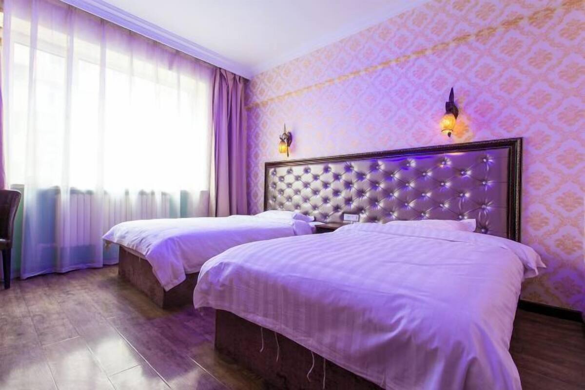 Een hotelkamer met twee bedden, een groot raam met gordijnen en behangpapier met een patroon.