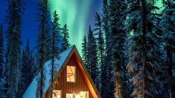 A wooden cabin with a triangular roof surrounded by snow-covered ground and tall evergreen trees.