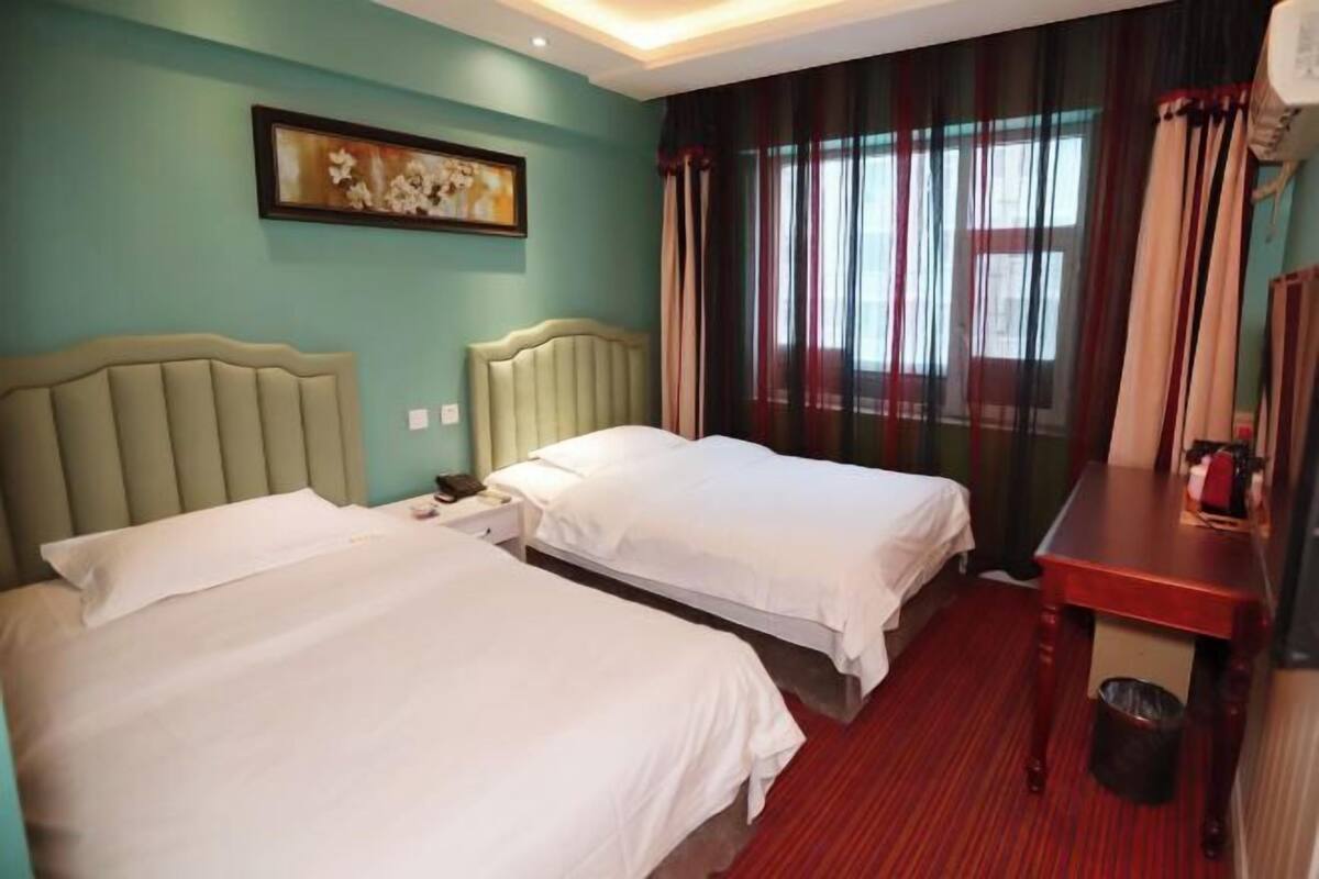 A hotel room with two beds, a desk, a trash bin, and a window with curtains.